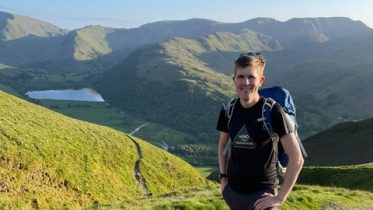 A picture of Alex, wearing a T-shirt with the Mind Over Mountains logo, standing in front of some green mountains.