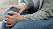 A person is seated on a couch, wearing a light gray sweater and blue jeans. They are holding their knee with both hands, indicating discomfort or pain in the knee area. The image focuses on the person's lower body and hands, with the face not visible.