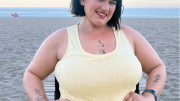 Image of Sophie Butler, seated in her wheelchair on a sandy beach. She has short black hair and is wearing a shimmering pale yellow sleeveless dress. The backdrop features a calm sea under a clear sky.