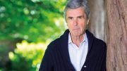 A portrait of an John Stapleton standing by a large tree in a what seems to be a park. He is wearing a navy blue cable-knit cardigan and a light blue collared shirt. He has his hands in his pockets as he leans against the tree.