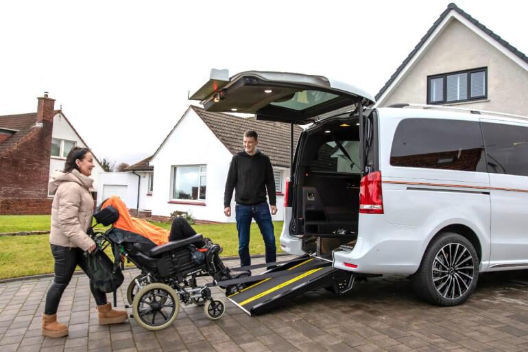 An image of a Brotherwood bespoke wheelchair accessible vehicle parked on a paved driveway in front of a plain house. A woman pushes someone in a wheelchair into the van using the fitted ramp.