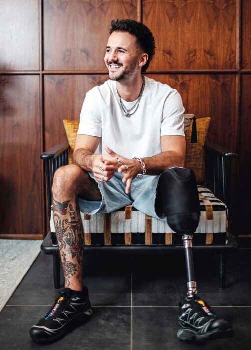 Luke Tarrant, a young man with a prosthetic leg and tattoos, sits in a stylish chair wearing a white T-shirt and denim shorts. He is smiling, hands clasped, and looking off to the side in a warmly lit room with wooden panelling in the background.