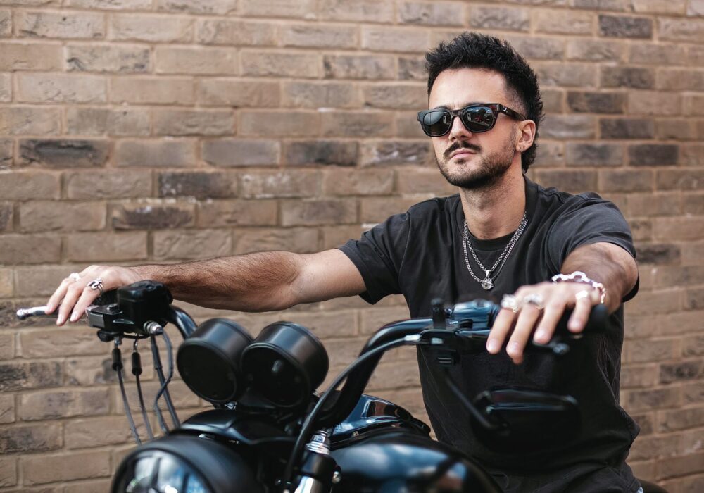 Luke Tarrant sits confidently on a black motorcycle, wearing sunglasses, a black T-shirt, and silver jewellery. His hands grip the handlebars as he looks off into the distance. There is a brick wall in the background.