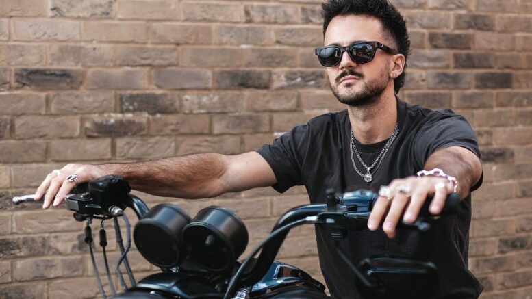 Luke Tarrant sits confidently on a black motorcycle, wearing sunglasses, a black T-shirt, and silver jewellery. His hands grip the handlebars as he looks off into the distance. There is a brick wall in the background.