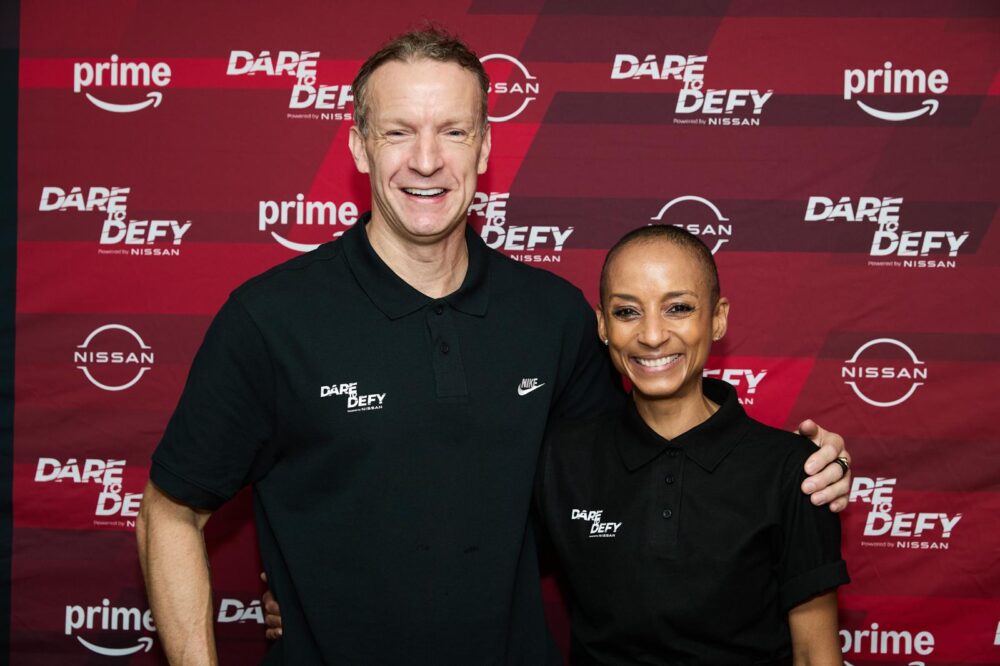 Richard Whitehead MBE and Adele Roberts stand side by side, smiling at the camera. They are wearing black polo shirts with "Dare to Defy" and Nike logos. They stand in front of a red banner with "Dare to Defy," "Nissan," and "Prime" branding.