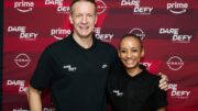 Richard Whitehead MBE and Adele Roberts stand side by side, smiling at the camera. They are wearing black polo shirts with "Dare to Defy" and Nike logos. They stand in front of a red banner with "Dare to Defy," "Nissan," and "Prime" branding.