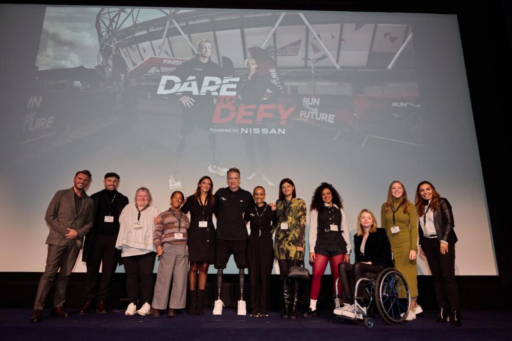 A group of people pose on a stage in front of a large screen displaying the "Dare to Defy" docu-series poster. They are dressed in a mix of casual and professional attire, all smiling for the photo.