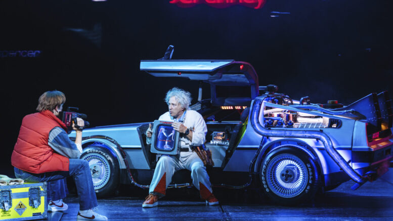 A stage performance of 'Back to the Future: The Musical' featuring two characters resembling Marty McFly and Doc Brown. Marty, wearing a red vest and jeans, is holding a video camera, while Doc, dressed in a white jumpsuit, is seated by the open door of a DeLorean time machine, holding a flux capacitor. The stage is illuminated with blue and neon lighting, and a "JCPenney" sign glows in the background, recreating an iconic moment from the film.