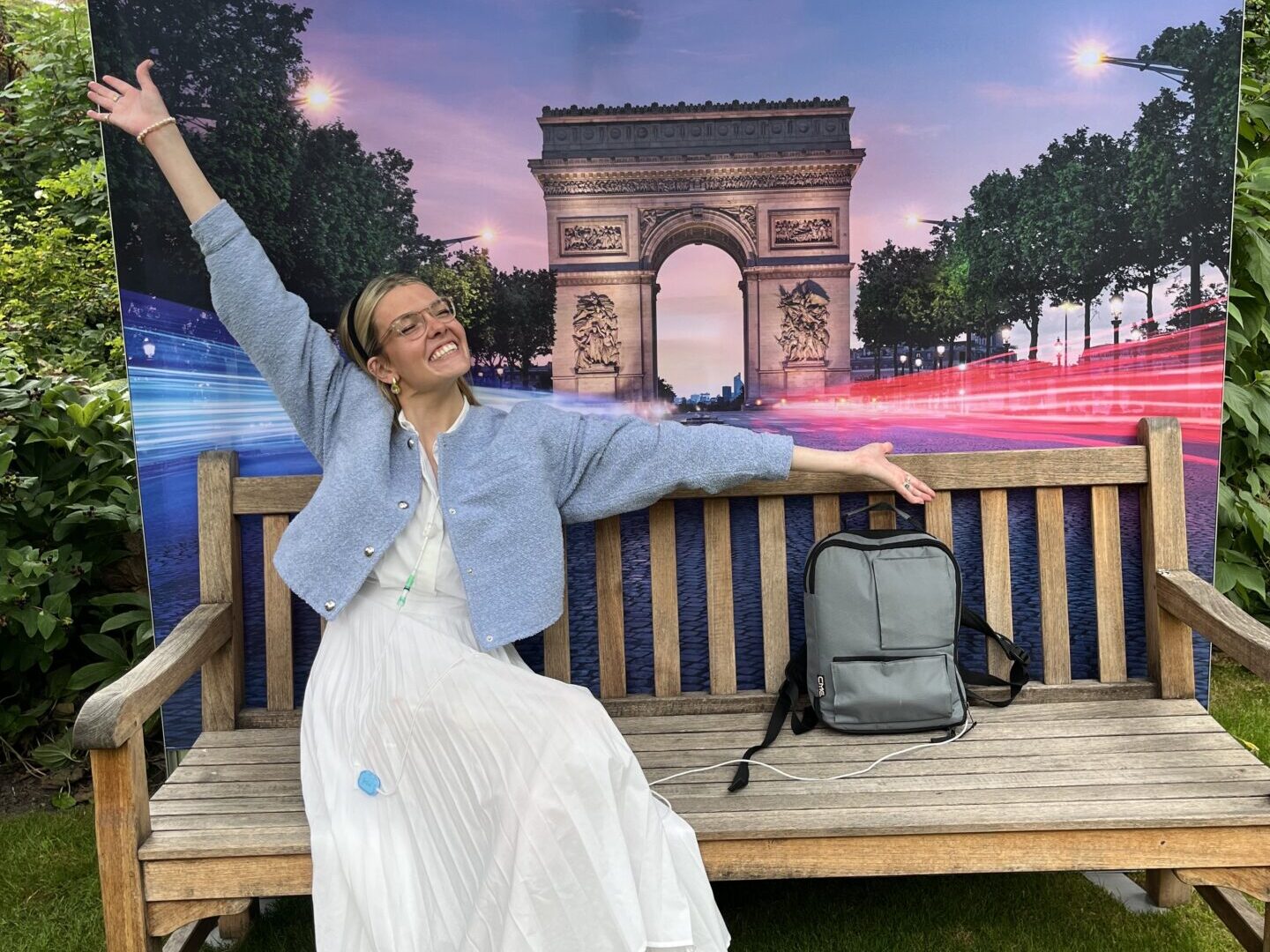 Celia sits on a wooden bench in front of a backdrop featuring the Arc de Triomphe and the logos of Channel 4 and the British Embassy in Paris. She wears a white dress, a blue cardigan, and sneakers, with a medical device visible on her side. She spreads her arms in a joyful pose, smiling brightly.