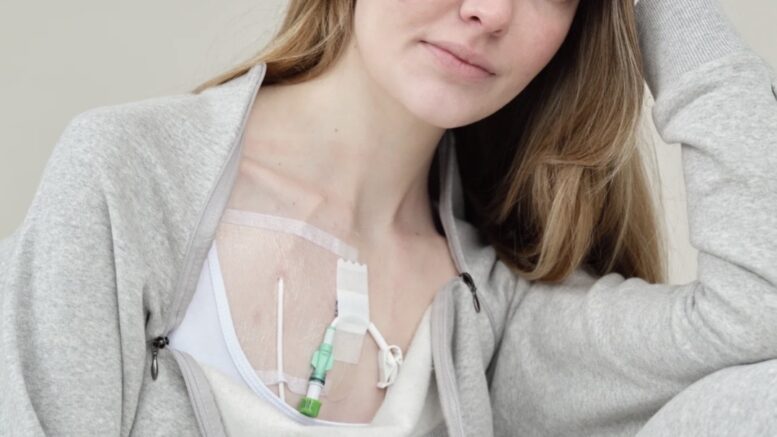 A photo of Celia who sits in a relaxed pose wearing a gray tracksuit. A medical port is visible on her chest. She gazes at the camera with a calm and confident expression.