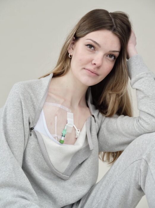 A photo of Celia who sits in a relaxed pose wearing a grey tracksuit. A medical port is visible on her chest. She gazes at the camera with a calm and confident expression.