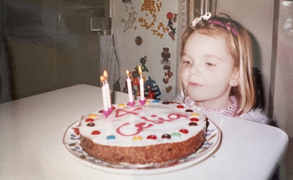 A picture of Celia as a child. She sits at a table in front of a birthday cake with three lit candles. The cake has colorful decorations and “Celia” written in icing. She is about to blow out the candles and looks excited.