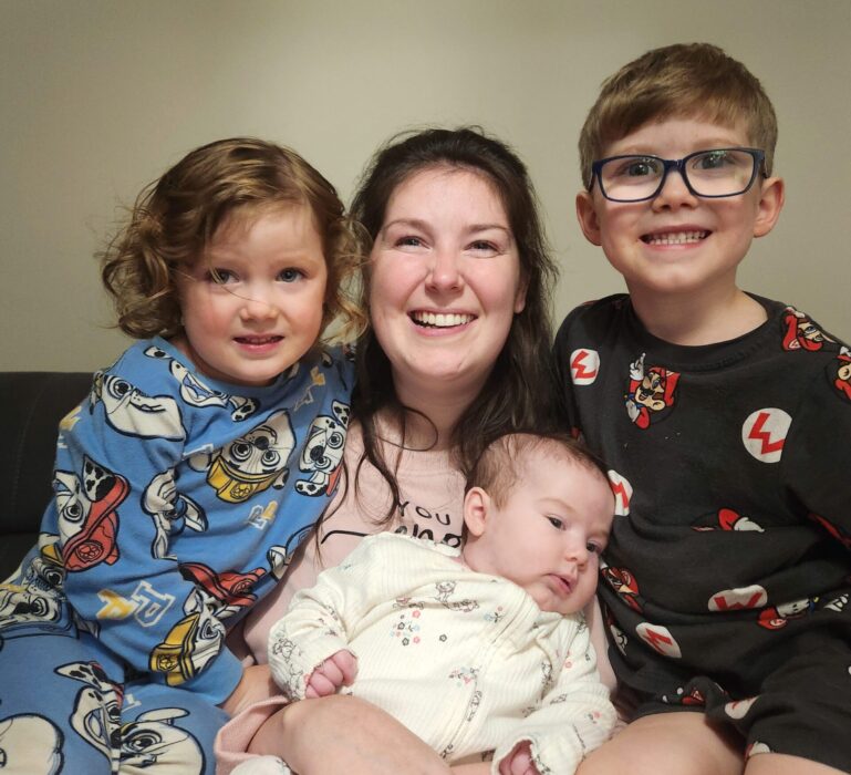 Lucy is sitting on a couch with her three kids, all cuddled up and smiling. She wears a cosy pink sweater. Her oldest daughter wears blue, Paw Patrol pyjamas and sits to her left, whilst her son wears black, Super Mario pyjamas and black glasses on her right. She holds her youngest, who is wearing a white, floral onesie, in her arms. 