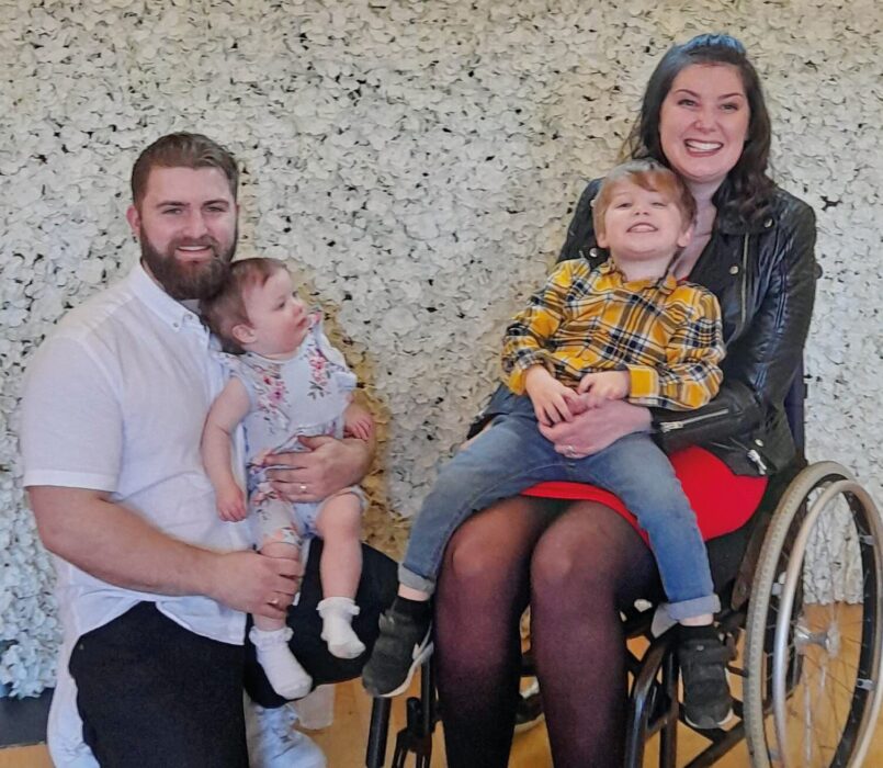 Lucy Lintott is sitting in her wheelchair, smiling while holding her young son on her lap. Next to her husband is kneeling and holding their baby daughter.  They’re in front of a pretty white floral backdrop. The family smiles happily at the camera. 