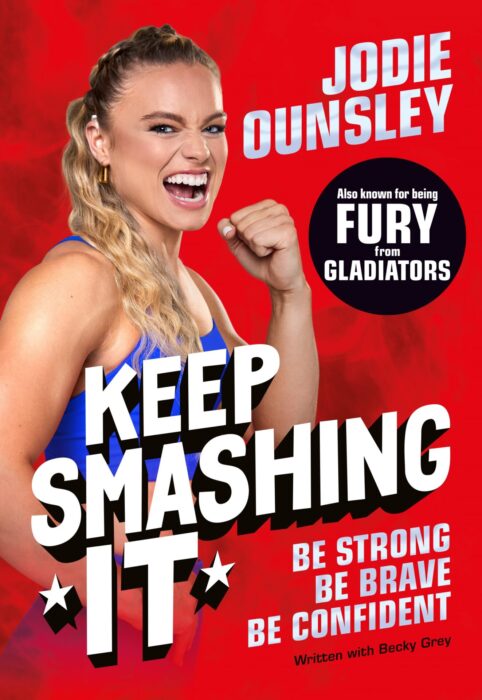 Jodie Ounsley, in a blue sports outfit, poses on the cover of her new book. She has a fierce expression and clenches her fist. The background is red. The book title "Keep Smashing It" is written in capital letters in bold, white text in the bottom left corner and her name is in the top right. There is a black circle with "Also known for being Fury from the Gladiators" in white text underneath her name. In the bottom right, smaller text in silver says "Be Strong, Be Brave, Be confident" in capital letters Underneath this is smaller black text which says "Written with Becky Grey."