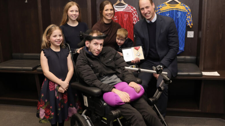 Rob Burrow sits in a wheelchair with his family around him in a locker room. His wife Lindsey and their three children stand beside him, all dressed in formal attire. Prince William, kneeling next to Rob, holds an award while smiling at the camera. Rugby jerseys hang in the background.