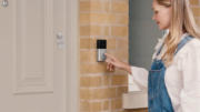 A woman wearing denim overalls and a white blouse is pressing an illuminated button on a smart video doorbell mounted on a brick wall beside a beige front door.