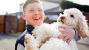 A smiling boy with Downs syndrome holds a fluffy white and tan dog, smiling brightly. They stand outside on a residential street. The background is out of focus.