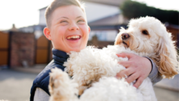 A smiling boy with Downs syndrome holds a fluffy white and tan dog, smiling brightly. They stand outside on a residential street. The background is out of focus.