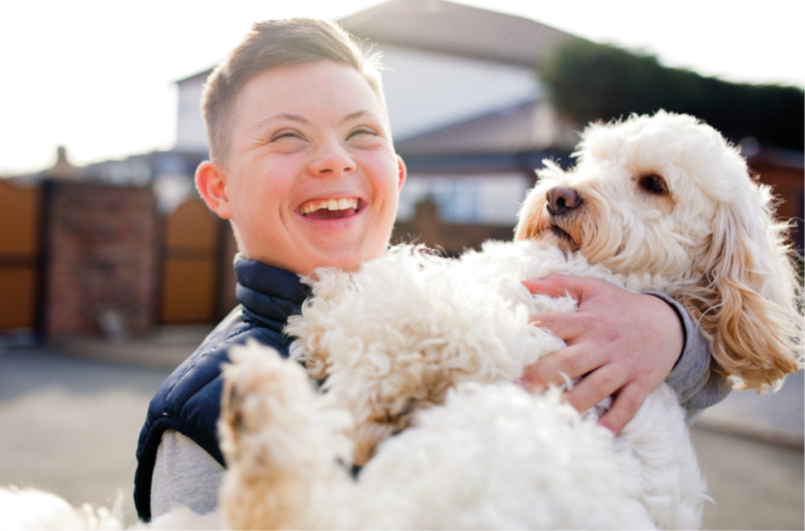 A smiling boy with Downs syndrome holds a fluffy white and tan dog, smiling brightly. They stand outside on a residential street. The background is out of focus.