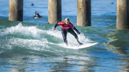 Zoe smith surfs a wave near some large pier pillars. She is wearing a wetsuit and a red t-shirt.