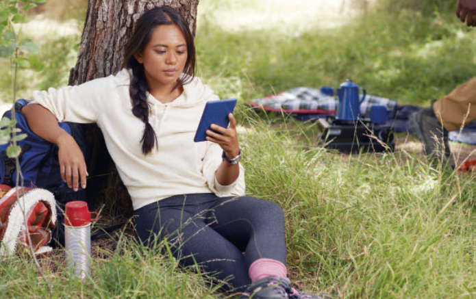 A young woman sitting under a tree, reading a kindle, with camping gear and grass around her.