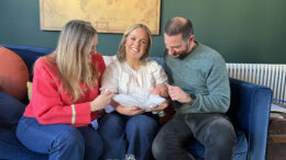 Ellie Simmonds sits on a blue sofa, smiling while holding a newborn baby. She is sat between two other adults -- a woman in a red sweater and a man in a green jumper -- who are lovingly admiring the baby. A vintage world map hangs on the green wall behind them.