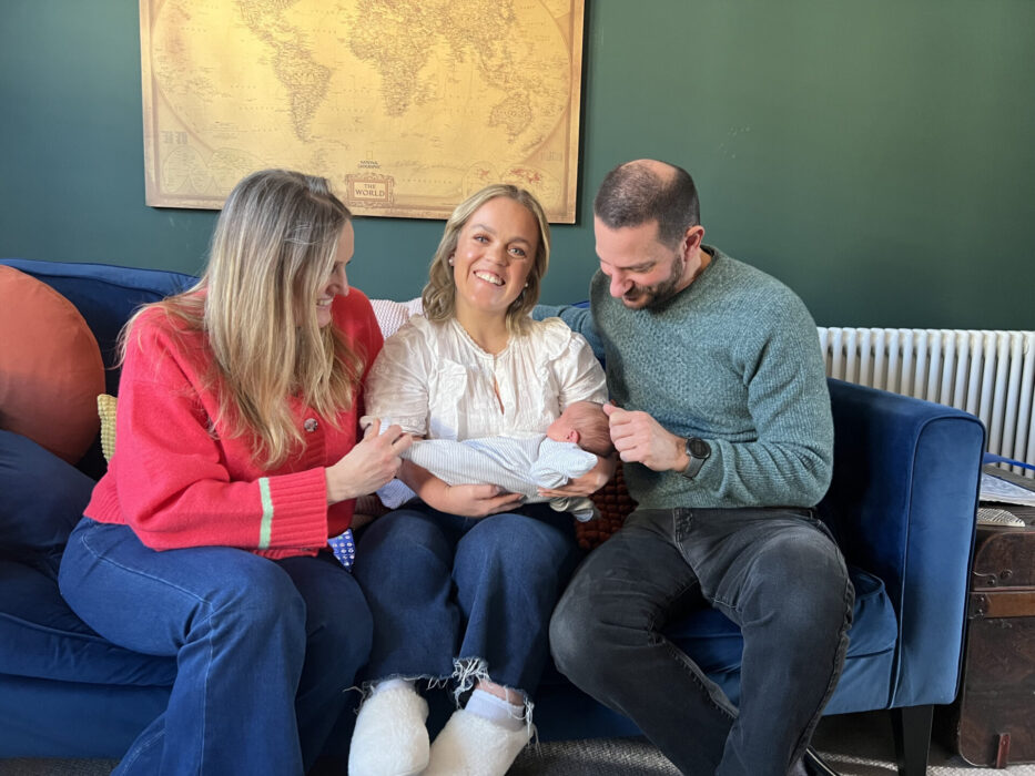 Ellie Simmonds sits on a blue sofa, smiling while holding a newborn baby. She is sat between two other adults -- a woman in a red sweater and a man in a green jumper -- who are lovingly admiring the baby. A vintage world map hangs on the green wall behind them.