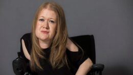 Frances Ryan, seated in a wheelchair, poses against a plain grey background. She has long, straight auburn hair and is wearing a black top with cut-out shoulders. She looks confidently at the camera.