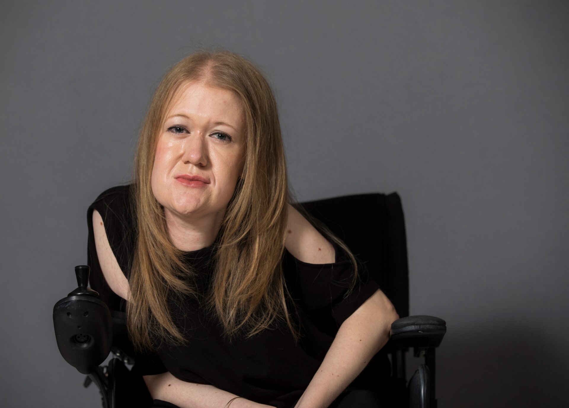 Frances Ryan, seated in a wheelchair, poses against a plain grey background. She has long, straight auburn hair and is wearing a black top with cut-out shoulders. She looks confidently at the camera.