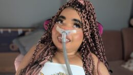 A photo of Shelby Lynch in her chair at home, wearing a white bandeau top and denim skirt. Her braided hair has pink highlights, and falls around her waist. She is using a breathing apparatus which is attached to the back of her chair. She smiles at the camera.