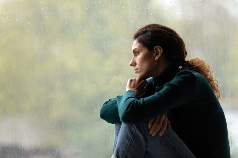 A woman in a green sweater and jeans sits by a rain-covered window, looking thoughtfully outside with her chin resting on her hands.