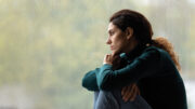 A woman in a green sweater and jeans sits by a rain-covered window, looking thoughtfully outside with her chin resting on her hands.