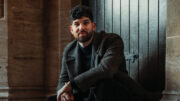 A picture of Mesha sitting on a stone step in front of a dark wooden door. He is wearing a dark patterned coat, black pants, and black boots.