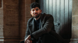 A picture of Mesha sitting on a stone step in front of a dark wooden door. He is wearing a dark patterned coat, black pants, and black boots.