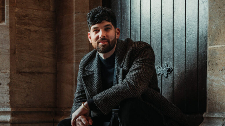 A picture of Mesha sitting on a stone step in front of a dark wooden door. He is wearing a dark patterned coat, black pants, and black boots.