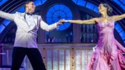 An image of a male and female theatre actor on stage dancing. They are styled in fancy costumes from the 1930s: the male is wearing a black and white tuxedo, while the women wears a purple dress with her hair pinned in an updo. They hold hands as they look at each other. The image captures them mid-dance in front of a speakeasy bar for the show Top hat.