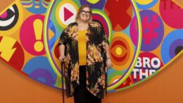 An image of Kerry Riches standing in front of the iconic Big Brother logo. She is smiling, wearing glasses, a yellow top, a floral black cover-up, and holding a walking stick.