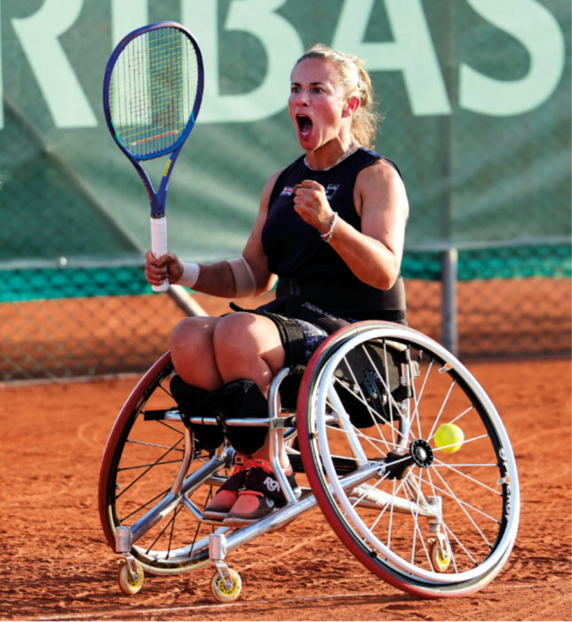Lucy Shuker, tennis icon, celebrates passionately during a match on a clay court. She clenches one of her fists and in the other hand holds a tennis racket. Her mouth is open wide as she cheers.  