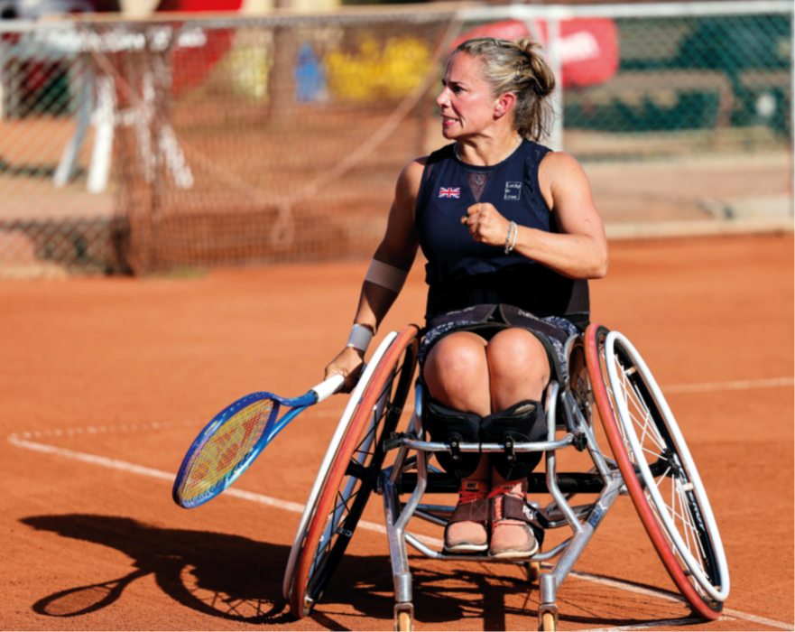 Lucy Shuker, a tennis icon, is pictured playing on a clay court. She clenches one of her fists and in the other hand holds a tennis racket. Her expression is determined, but focused. 