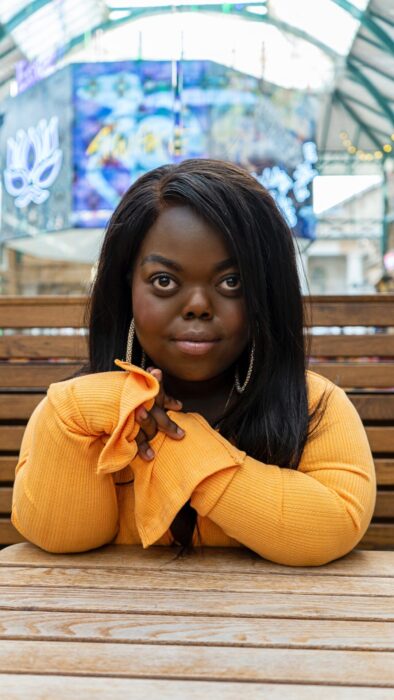 The image shows Fats Timbo sitting at a wooden table, wearing a bright orange long-sleeve top with flared cuffs. She has long dark hair and is looking directly at the camera with her hands gently clasped in front of her. The background appears to be an indoor market or shopping area with colourful lights and signs.