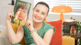Molly Burke sits on a yellow couch, smiling and holding up her new memoir "Unseen: How I Lost My Vision but Found My Voice." She wears a green outfit with gold and beaded bracelets. She appears to be a living room. Behind her is a stack of her books, a bright orange lamp, and a window with white blinds.