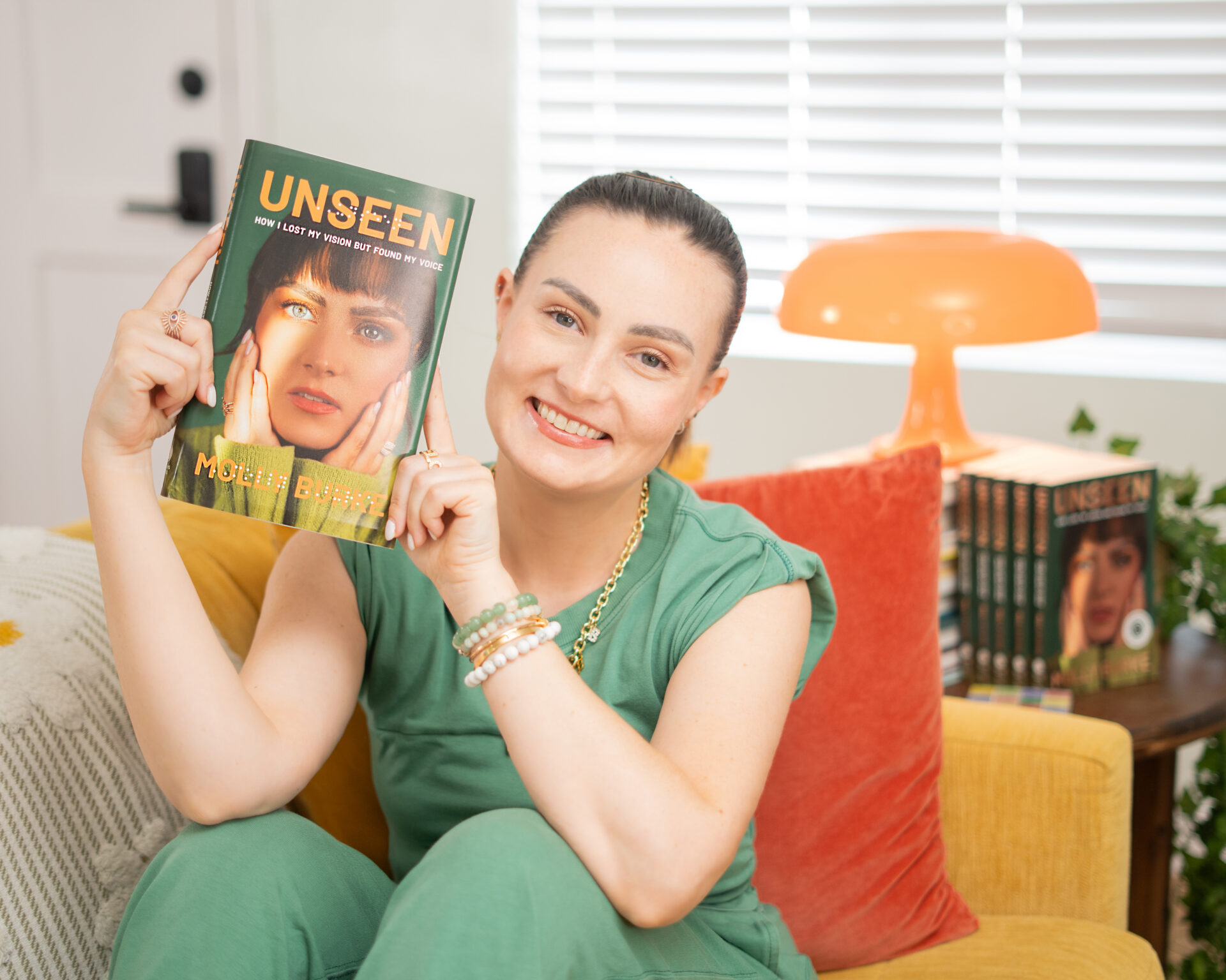 Molly Burke sits on a yellow couch, smiling and holding up her new memoir "Unseen: How I Lost My Vision but Found My Voice." She wears a green outfit with gold and beaded bracelets. She appears to be a living room. Behind her is a stack of her books, a bright orange lamp, and a window with white blinds.