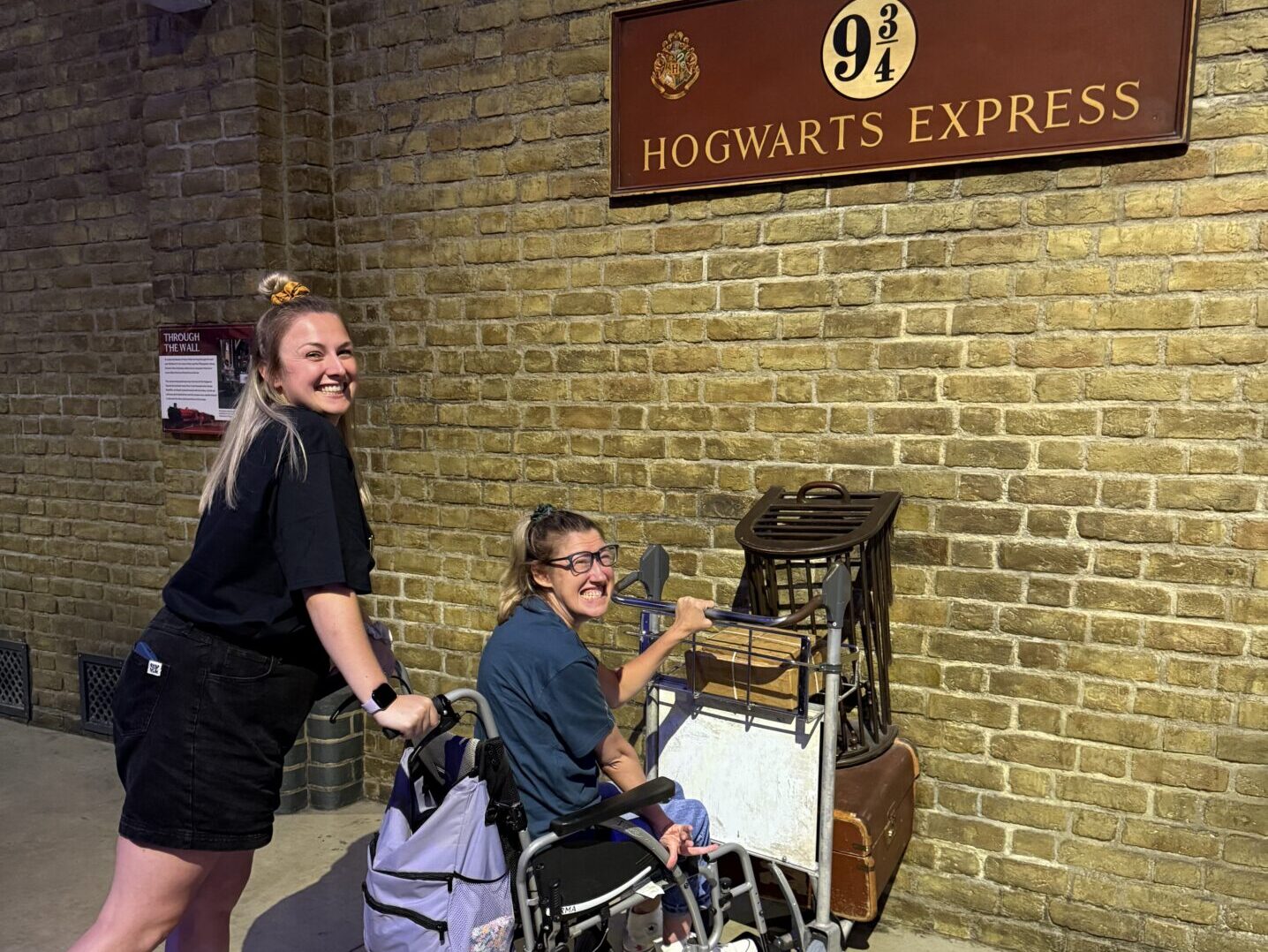 Hannah and Becky Cheetham at the Warner Bros. Studio Tour London -The Making of Harry Potter. They are pictured in front of the platform 9 and three quarters sign. Becky is pushing Hannah in her chair, who is holding onto the famous trolley prop which is halfway through the wall. 