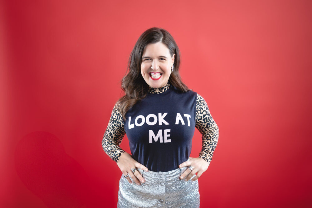 Rosie Jones standing against a bold red background, grinning, and wearing a “LOOK AT ME” top with leopard print sleeves and a silver skirt.