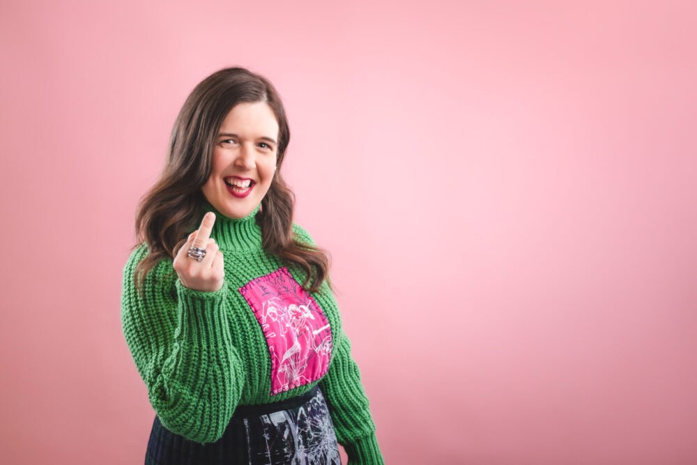 Rosie Jones smiling in front of a pink background, wearing a green jumper and holding up her hand with a cheeky expression.