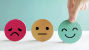 Graphic showing three round faces representing emotions. From left to right: a pink sad face with a frown, an orange neutral face, and a green happy face being picked up by a hand.