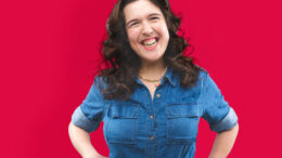 Rosie Jones stands laughing in front of a bright red background. She's wearing a denim shirt with her hands on her hips. She's visible from the waist up.
