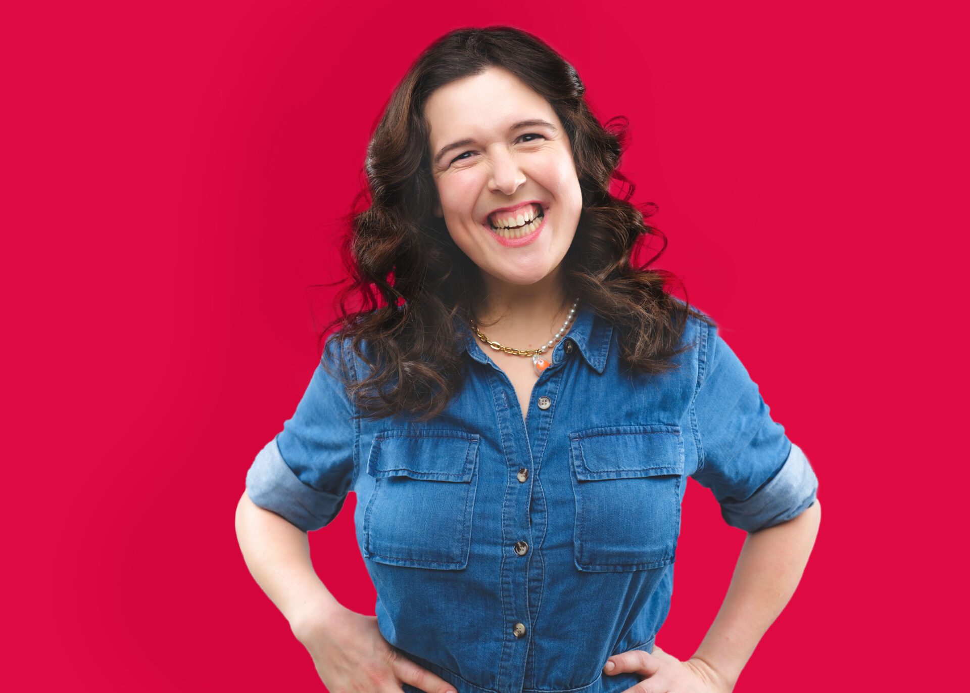 Rosie Jones stands laughing in front of a bright red background. She's wearing a denim shirt with her hands on her hips. She's visible from the waist up.