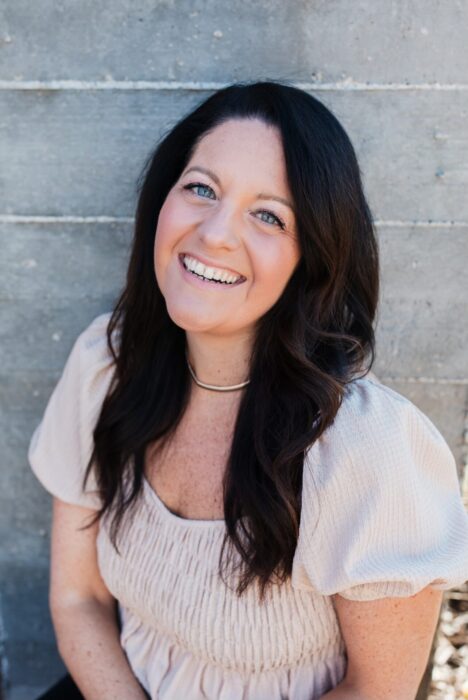 Portrait photo of Alison Seponara, a smiling woman with long dark hair, wearing a light beige blouse. She is sitting in front of a gray cement wall and looking directly at the camera.